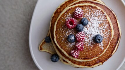 Top-down view of a plate of pancakes with berries and powdered sugar, styled for a cooking video, highlighting a rustic and cozy breakfast theme. - Powered by Adobe