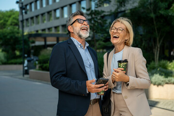 Happy mature businessman and businesswoman laughing and chatting outdoors during coffee break in corporate setting