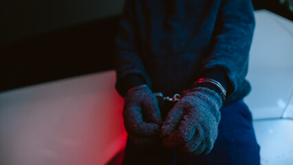 Handcuffed individual wearing gloves, sitting on a surface in a dimly lit setting with colorful...