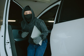 Person in dark clothing sneaking out of car holding laptop in a dimly lit parking garage during...