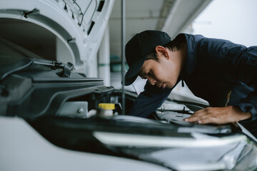Mechanic inspecting car engine during cloudy day in parking garage while using laptop for...