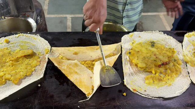 Close-up of Indian street food vendor spreading spicy potato filling on dosa over hot tawa showcasing traditional South Indian cooking preparation process in kitchen setup