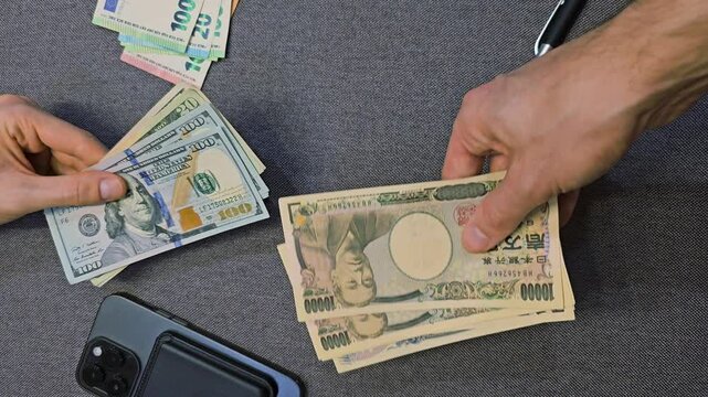 Hands exchanging US dollars and Japanese yen banknotes with euro cash on table near smartphone, international currency conversion and financial transaction concept, top view close up