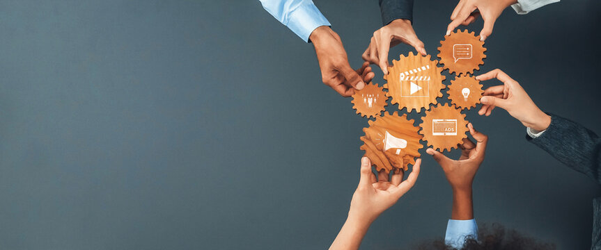 This image portrays a vibrant teamwork scenario where diverse hands hold wooden gears adorned with icons, representing collaboration and innovative business strategies. Amity