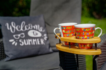 Scene with two coffee cups in a special wooden cup holder, a chair and a cushion in a garden.