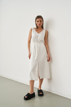 Woman in sleeveless white dress with V-neck and black shoes stands relaxed, leaning against a wall, with a neutral expression, hands by her side, on a light-colored floor