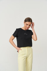 Woman in black shirt with subtle text, wearing pale yellow pants, gently touching her hair. Her...