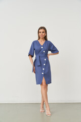 A woman wearing a blue dress with white buttons stands confidently with one hand on her hip. Her hair is long and brown. The dress has short sleeves and a belt tied at the waist