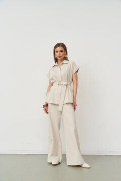 Woman wearing a beige belted top and wide-leg trousers, standing confidently with hands at her side. White shoes complement the minimalist, chic outfit in a neutral setting