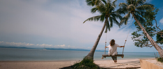 A young girl swinging on a swing between palm trees on the beach of a tropical island. Young woman watching the ocean on a sunny day