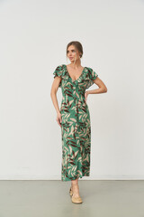 Woman wearing a long green dress with a tropical leaf pattern. Her hands are on her hips, and she...