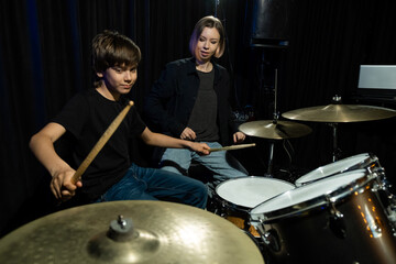 Young caucasian woman teaches a boy to play the drums in the studio on a black background. Music...