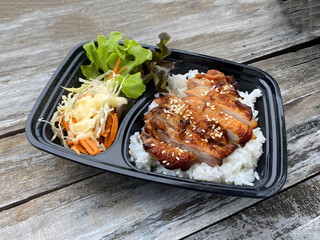 Grilled chicken served on steamed rice, garnished with sesame seeds, accompanied by fresh lettuce, shredded cabbage, and carrot salad with mayonnaise dressing. Presented in a black takeaway box on a r
