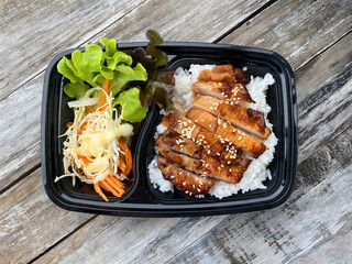 Grilled chicken served on steamed rice, garnished with sesame seeds, accompanied by fresh lettuce, shredded cabbage, and carrot salad with mayonnaise dressing. Presented in a black takeaway box on a r