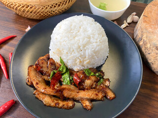Delicious Thai grilled pork neck stir-fried with chili and holy basil served with steamed jasmine rice. Traditional Thai street food presented on a black plate with fresh red chili and garlic in the b