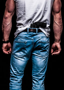 A man is seen from the back, wearing jeans and a white t-shirt. A handgun is tucked into the waistband of his jeans. The man's arms are muscular and his fists are clenched.