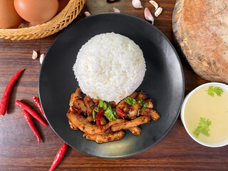 Delicious Thai grilled pork neck stir-fried with chili and holy basil served with steamed jasmine rice. Traditional Thai street food presented on a black plate with fresh red chili and garlic in the b