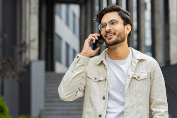 Happy Indian man having remote conversation communicate speaking by smartphone outdoors. Arabian Hindu guy talking on phone unexpected good news gossip walking in downtown city street. Business people
