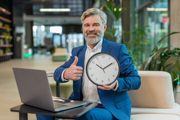 It is your time. Mature boss businessman with laptop showing time on wall clock, approve, pointing...