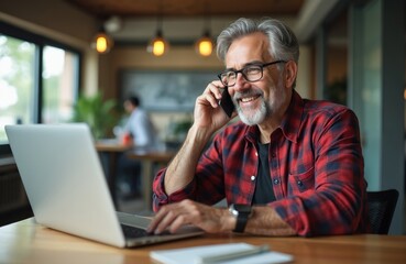 Smiling grey-haired man talks on phone near laptop at home office. Happy male working, using computer technology, mobile phone. Casual business attire. Video call, remote work, telework, cyberspace