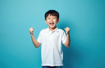 Joyful asian little boy wearing white t-shirt celebrates victory. Korean child with smile in excitement against blue background. Happiness fun. Studio portrait.