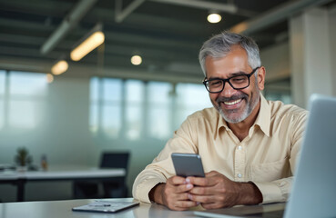 Smiling middle-aged Hispanic businessman CEO using cell phone in office. Mature Latin or Indian man works on mobile app, laptop. Happy man working online, connects with clients, partners. Online job.