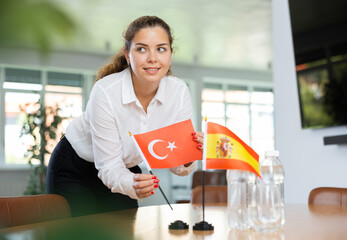 Positive young woman putting little flag of Turkey on table next to the flag of Spain and bottles...