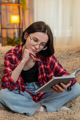 Obraz premium Young woman sits on home sofa, reading through old personal journal notebook and smiling with nostalgic mood. Girl reflects on memories with calm expression, embracing past thoughts and quiet emotion.
