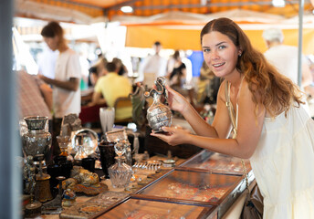 Girl in flea market store look at silver decanter, antique gifts. Female buyer holds item in hands...