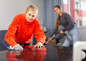 Portrait of positive european woman wiping the table at home, her daughter cleaning sofa with...