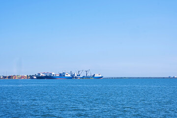 Cargo ships docked at port. Industrial seascape under clear sky. A wide shot across sea captures...