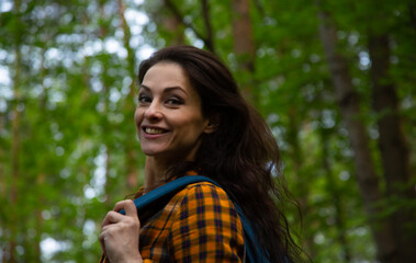Beauty face happy smiling woman looking on a green pine trees in the forest on sun spring weather. Girl walking in woods with eyes look front with backpack on shoulders. Closeup portrait