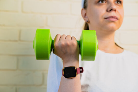 A woman lifts a green dumbbell with her right arm, while wearing a smartwatch and a white shirt.