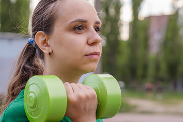 A woman in a green t-shirt performs a dumbbell shoulder press exercise during an outdoor workout...