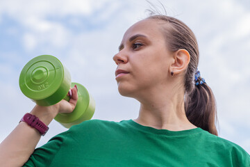 A woman in a green t-shirt performs a dumbbell shoulder press exercise during an outdoor workout...