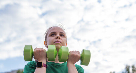 A woman in a green t-shirt performs a dumbbell shoulder press exercise during an outdoor workout...