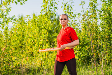 A woman is using a weighted hula hoop while wearing a red shirt and black pants. She is in a green...