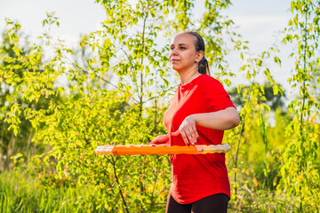 A woman is using a weighted hula hoop while wearing a red shirt and black pants. She is in a green...