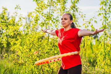 A woman is using a weighted hula hoop while wearing a red shirt and black pants. She is in a green...