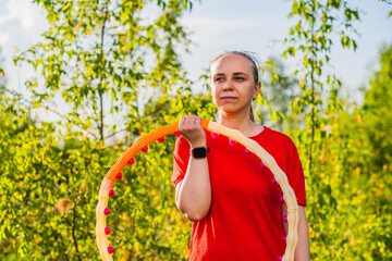A woman is using a weighted hula hoop while wearing a red shirt and black pants. She is in a green...