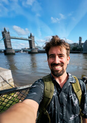 Vertical. Happy male tourist with backpack taking a selfie with tower bridge in the background, enjoying summer holidays in london, united kingdom, with city hall visible on the right