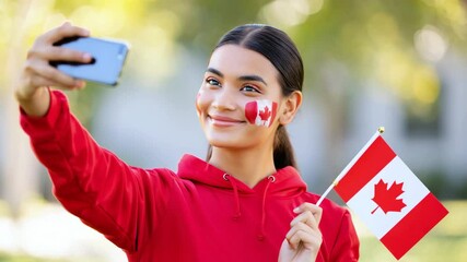 Young woman in red hoodie taking selfie with smartphone, holding Canadian flag and face painted with maple leaf outdoors in daylight, Canada Day, national pride, celebration