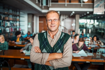 Portrait of a confident male professor standing in a library with students studying in the background
