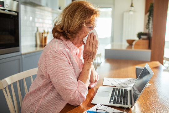 Senior woman sneezing in tissue while working on laptop at home - Powered by Adobe