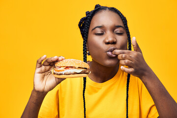 Young woman enjoying a delicious hamburger with a playful expression, vibrant yellow background,...