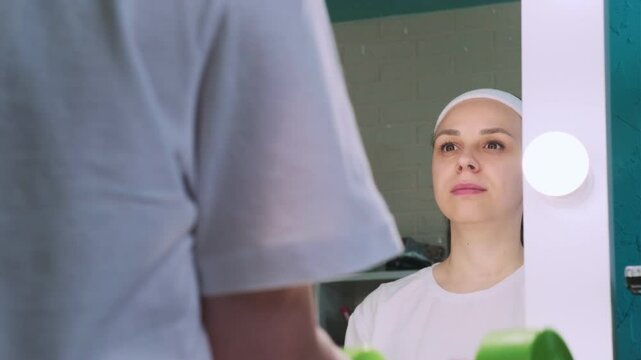 A woman in a white shirt and a headband lifts weights while looking at herself in a vanity mirror. The mirror has a row of lights around the frame.