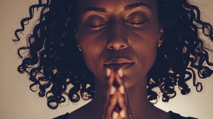 Close-up video still of a woman meditating with eyes closed, hands in prayer. Warm lighting, soft focus, and a serene, introspective mood. - Powered by Adobe