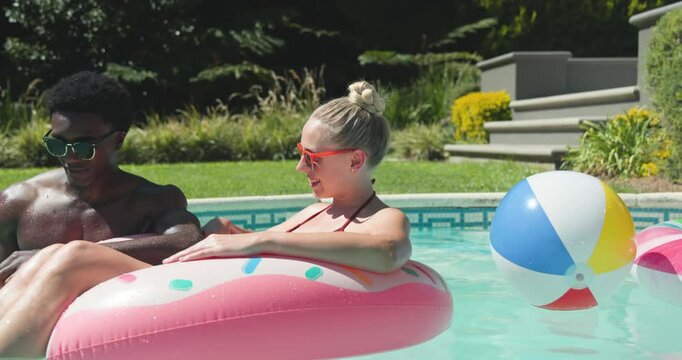 Diverse couple bantering with man splashing smartphone as woman lounging on pink float in pool