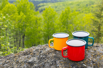 Three metallic tea cups—red, blue, and yellow—on ancient mountain rocks , set against a breathtaking view of a forested valley in sun light. Camping and hiking vibes of adventure.