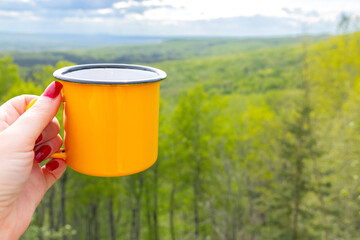 A woman's hiker hand holding a bright yellow metallic cup on the top of a mountain in front of breathtaking view of a forested valley in sun light. Camping and hiking vibes of adventure.